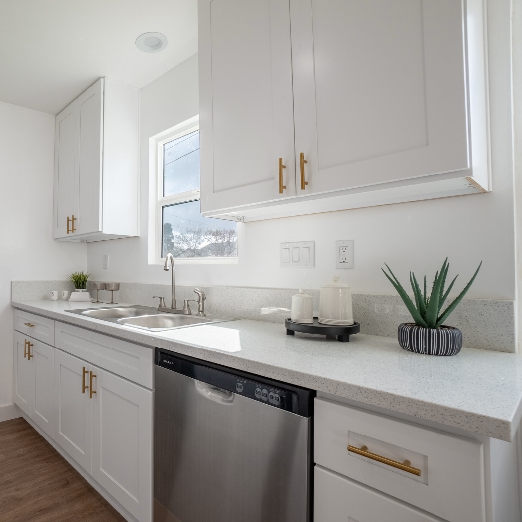 A modern kitchen in an ADU featuring sleek white cabinetry with brass handles, a stainless steel dishwasher, and a polished quartz countertop. The space is well-lit with natural light streaming in through a window above the double sink, highlighting the minimalist design and high-end finishes. A small potted plant adds a touch of greenery to the clean and elegant setting.