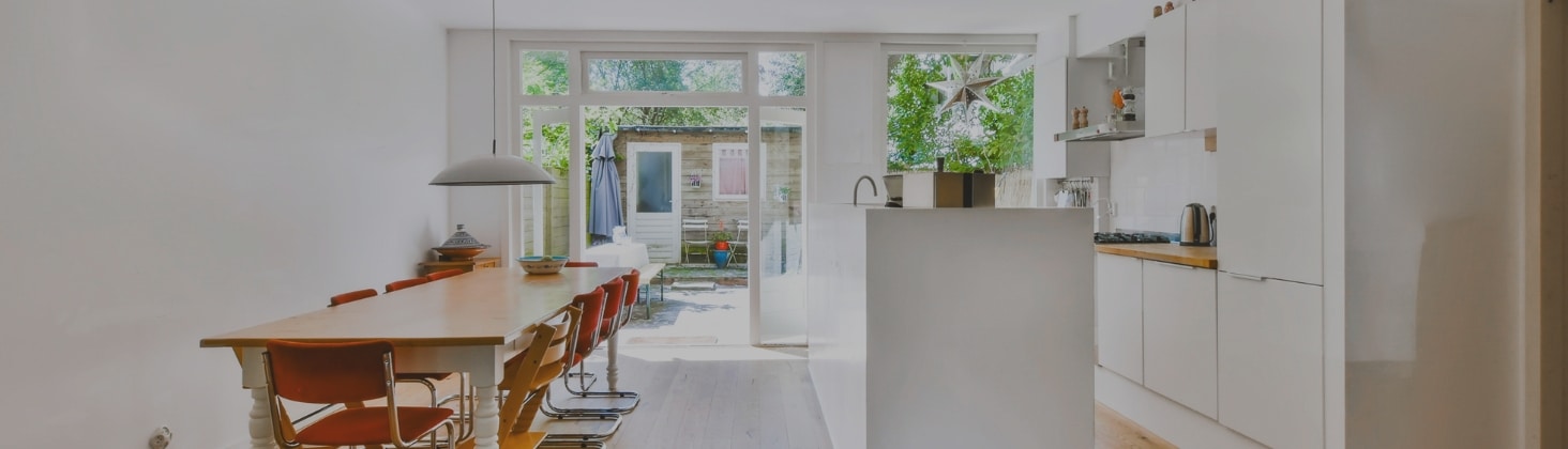 Bright and airy garage conversion ADU featuring a minimalist kitchen with white cabinetry and countertops, a long wooden dining table with red chairs, and large glass doors opening to a serene outdoor space.