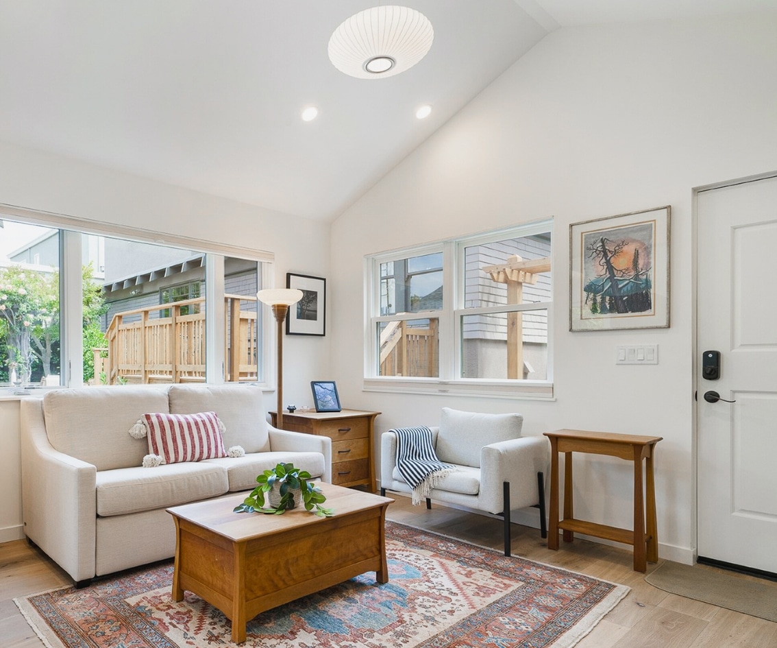 A cozy living space with a vaulted ceiling and ample natural light, featuring large windows that overlook a wooden deck outside. The room is tastefully decorated with a beige sofa adorned with a striped pillow, a matching armchair, a wooden coffee table, and a patterned rug. A floor lamp and framed artwork add to the inviting ambiance, making it a perfect example of a well-designed room addition by a skilled room addition contractor.